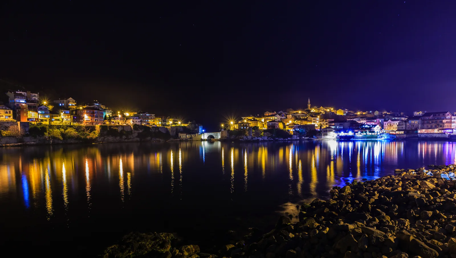 Gece taşların ardından görünen liman ışıkları ve suya vuran yansımaları